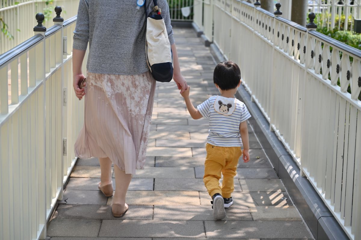 手をつないで並木の遊歩道を歩く母と幼児の後ろ姿。療育へ送っていく朝の場面